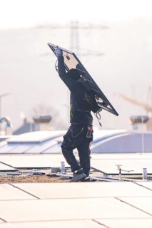 Worker in protective clothing lifts solar module on a sunny roof surrounded by urban landscape, PV