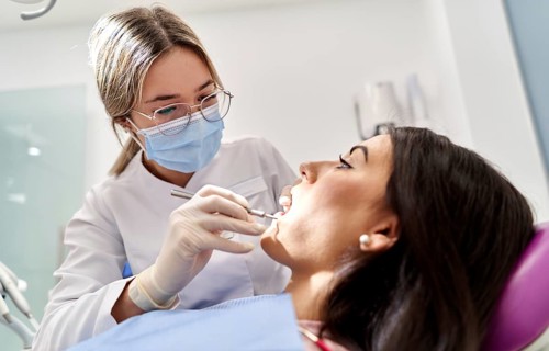 A dentist wearing a mask and gloves examines a patient's teeth, using tools in a modern dental