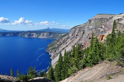 Steep rocks above the deep blue lake with green forest and expansive views of mountains on the