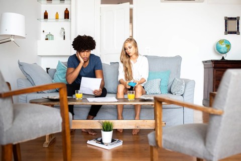 A couple sits on a sofa analyzing financial documents and using a calculator. The setting is a