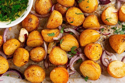 Roasted potatoes sit on a baking sheet next to chopped herbs and red onions. They are golden brown