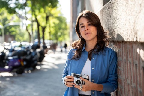 A stylish lesbian woman with wavy brown hair leans against a brick wall, holding a vintage camera.