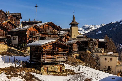 Snow-covered roofs of old farmhouses and barn storage, in the back the wooden cross and the church