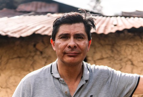 A close-up portrait of a man standing outdoors in a rural area of Colombia, with a rustic clay wall