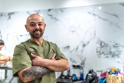 Smiling bald man with a beard and arm tattoo standing confidently with arms crossed, wearing a