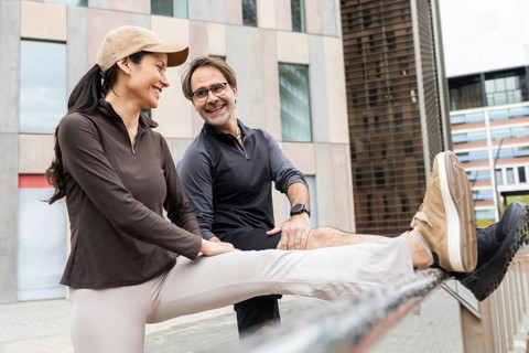 A couple smiles as they engage in an outdoor fitness routine, stretching against a railing. They