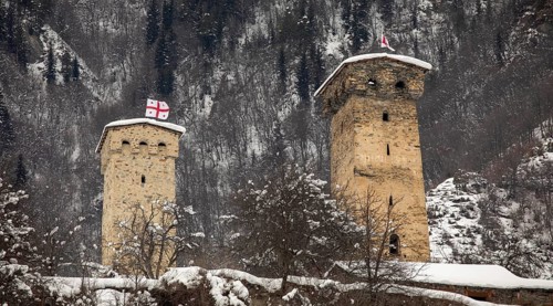 Two ancient stone defense towers stand amid a snowy landscape, with a forested mountain in the