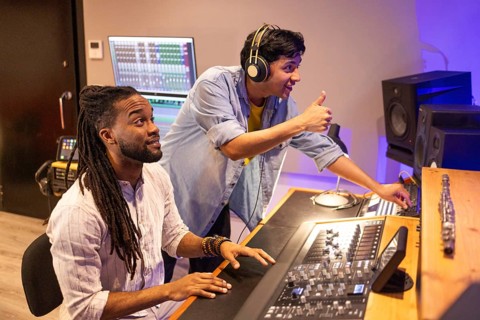 A vibrant recording studio scene showing two focused producers working on audio mixing. One uses a