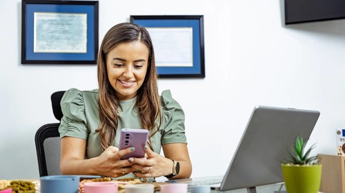 A nutritionist sits at her desk, smiling while using a smartphone, with a laptop open. Certificates