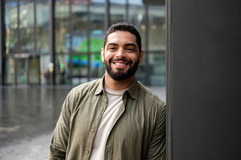 A confident man smiles warmly, wearing casual attire, while leaning against a wall in front of a