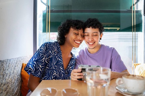 A joyful multiethnic LGBTQ+ couple smiles while sharing a tender moment at a cozy cafe browsing