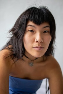 A close-up portrait of a young Asian woman with dark hair, bangs, and facial piercings. She wears a