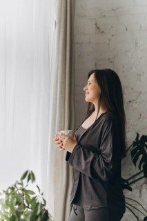 A woman stands by a window with a cup of coffee, enjoying the morning sunlight. Her serene