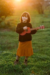 A young girl stands in a sunlit field, playing a ukulele. She wears a cozy sweater and mustard