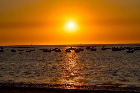 Several boats are silhouetted against a vibrant orange sunset over the ocean in sanlucar de