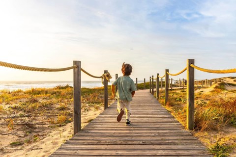 Young boy running along a wooden boardwalk through sand dunes toward the atlantic at golden hour,