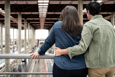 A loving lesbian couple embraces while standing on a balcony, overlooking a train station. Their