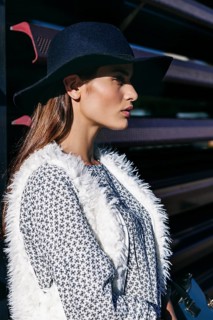Woman in a black hat and white fur vest poses stylishly. Her patterned dress adds elegance, while