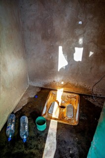 A shadowy bathroom with a simple toilet, two water bottles and a cup. Light falls through a window,