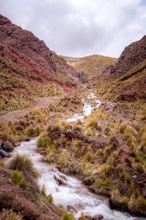 Scenic view of the Peruvian Andes with red soil, lush vegetation, and a winding stream. Dramatic