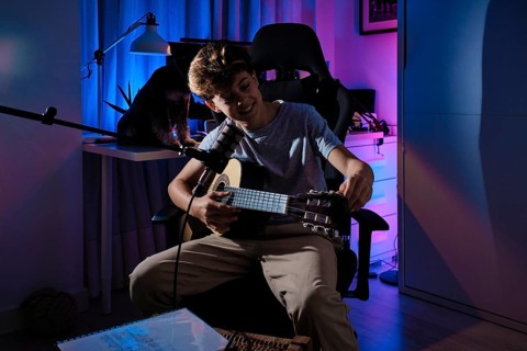 A young musician tunes a guitar in a cozy home studio, illuminated by colorful LED lights. The