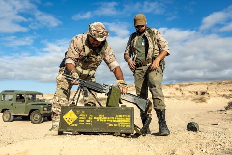 Two soldiers in camouflaged uniforms organize equipment next to a sand dune during a military