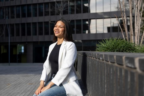 A confident woman wearing a white blazer sits outdoors, smiling in an urban setting. The modern
