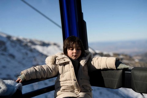 A child in a beige coat rests serenely inside a ski lift, surrounded by snowy peaks at Sierra