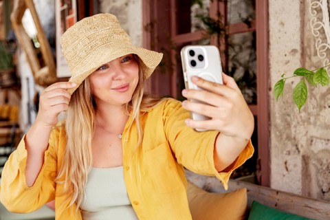 A young, cheerful woman in a yellow shirt and stylish hat takes a selfie with her mobile phone at a