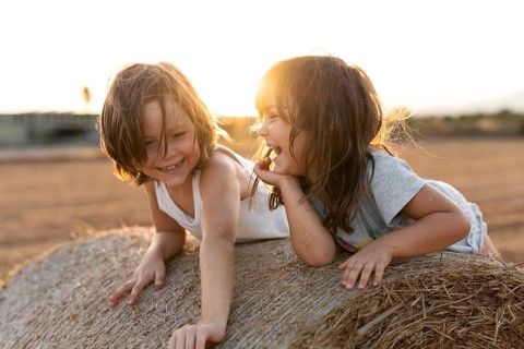 Two cheerful children are laughing and playing on a hay bale in a sunlit field, surrounded by