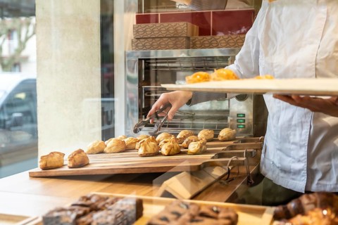 Baker's arms arranging freshly baked empanadas with tongs onto a wooden display shelf in a bustling