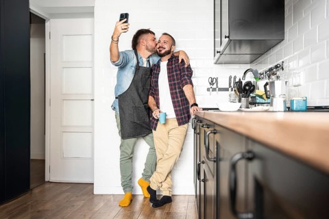 A joyful gay couple poses for a selfie in their modern kitchen. One partner embraces the other,