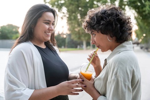 A lesbian couple sharing a joyful moment outside, with one partner sipping a fruit drink. The