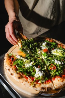 A person grabs a slice of pizza topped with fresh arugula and creamy cheese. The pizza is placed on