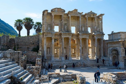 Ancient ruins with a well-preserved façade, palm trees in the background, visitors on the grounds,