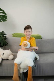 A young boy in a yellow shirt sits on a gray sofa, hugging a large plush goose toy. Nearby, a green