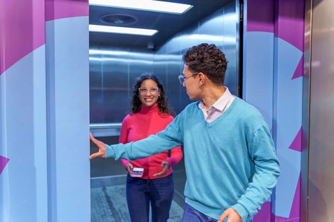 A Gen Z man holds the elevator door open for a happy woman holding a smartphone. They are casually