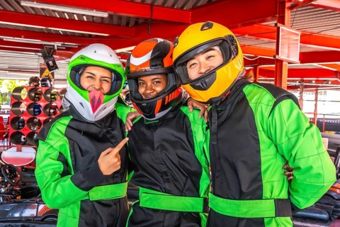 Three smiling diverse friends wearing helmets and racing suits, standing closely together at a