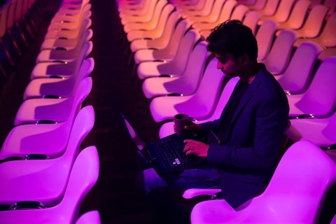 A indian man sits alone in an auditorium with purple lighting at a conference of AI in Amsterdam,