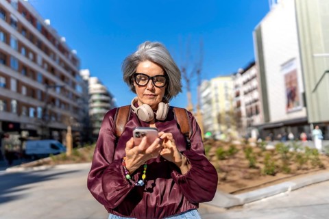 Woman with grey hair and headphones around her neck focusing on her smartphone screen while walking