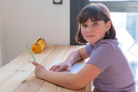 A young girl engaged in her homework sits at a wooden table, sporting a casual purple top and a