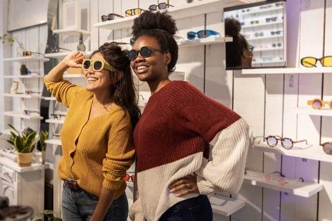 Two friends happily explore a stylish sunglasses store, trying on various pairs while surrounded by