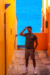Young man holding sunglasses while standing in a colorful alleyway with the blue ocean in the