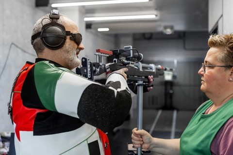 A blind athlete, wearing specialized gear, participates in a shooting competition. Guided by an