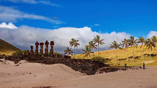 Stone statues on a sandy area of Easter Island, with palm trees in the background and blue sky, The