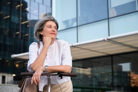 A mature woman with gray hair and a thoughtful expression leans on a scooter handle in front of a