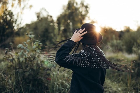 A woman stands with her back to the camera, touching her hat, as the sun sets behind her, casting a