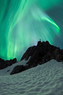Unrecognizable person silhouette stands atop a snow-covered mountain under the vibrant green swirls