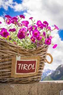 A basket filled with purple flowers and a Tyrol sign is placed in front of a mountain landscape,
