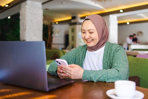 A Muslim woman wearing a hijab is smiling while using her smartphone and laptop at a cozy cafe. The